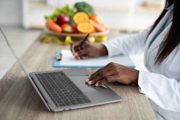 African american dietitian creating individual diet plan for patient or counting calories, working on laptop and clipboard, selective focus, closeup